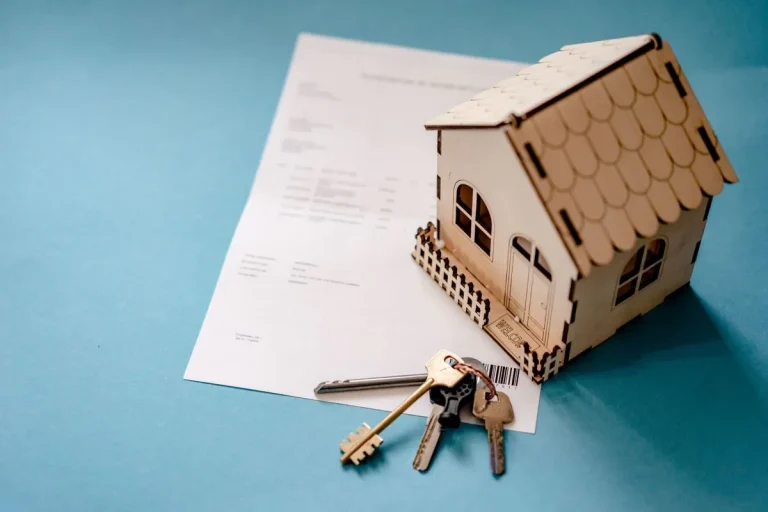Wooden model house and keys resting on paperwork on a blue table